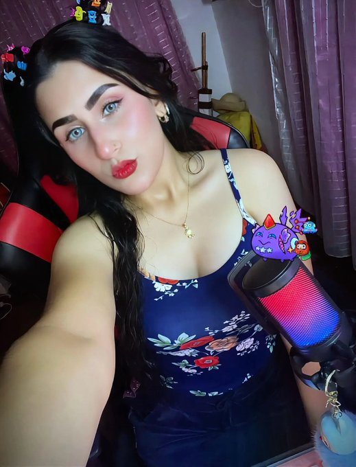 The image features a woman with blue eyes wearing a floral top and holding a microphone. She has a big smile on her face as she takes a selfie, which includes a caption about her day that reads "Good morning, big guys Today we woke up feeling super blue and very flowery, but with positive energy cranked up to the max always Come on, we're already starting, we're ready to enjoy the Sands of @AxieInfinity , we're gonna have a blast twitch.tv/kar3ncit4x". The woman's facial expression and body language convey her excitement about the day ahead.