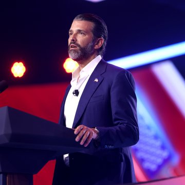 Donald Trump Jr. stands at a podium on a stage illuminated by red and blue lighting, an image paired with news that his venture firm 1789 Capital is leading a $1 billion funding round for Polymarket, valuing the prediction market platform at $21 billion.