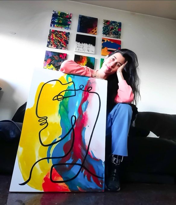 A young woman sits on a black couch beside a large abstract painting featuring a continuous black line over vibrant splashes of yellow, blue, red, and green, with a grid of similar smaller artworks hanging on the wall behind her.