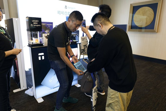 The image shows a group of people gathered around an exhibit at a convention or event, with one man holding up his phone for all to see. The background includes a large painting and a sign that reads "Luxury".