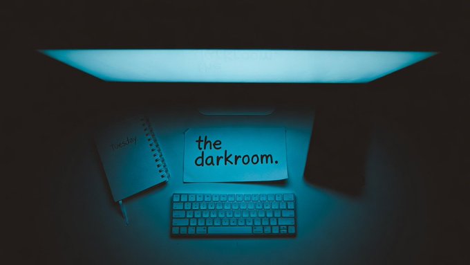 The image shows a computer setup with a monitor, keyboard, and mouse. The monitor displays the words "the darkroom" along with some text about a project or task that has been completed. A notebook and pen are also present on the desk next to the monitor.