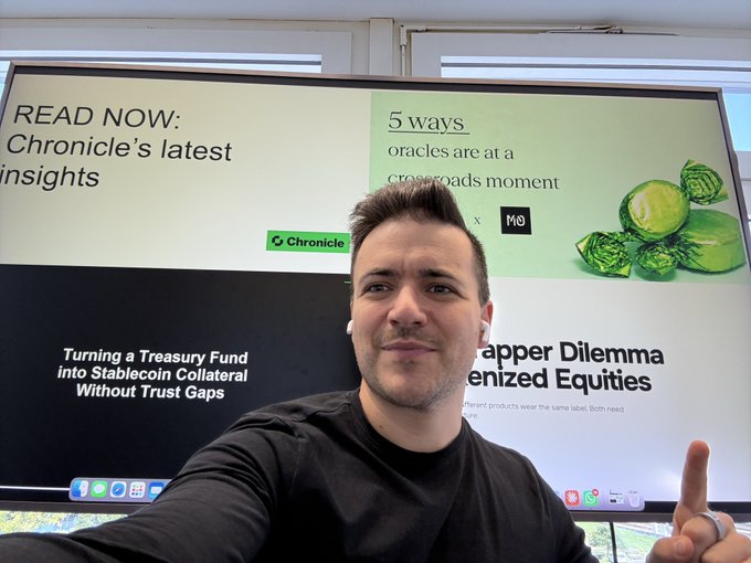 A man taking a selfie in front of a large display screen promoting Chronicle's latest insights, with visible text reading '5 Ways oracles are at a crossroads moment' and 'Turning a Treasury Fund into Stablecoin Collateral Without Trust Gaps'.