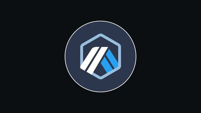 A dark circular icon featuring the Arbitrum logo—a stylized "A" composed of white and blue geometric shapes within a hexagonal frame—displayed on a gray background, representing Arbitrum's Layer 2 scaling solution and its new resource-based pricing model.