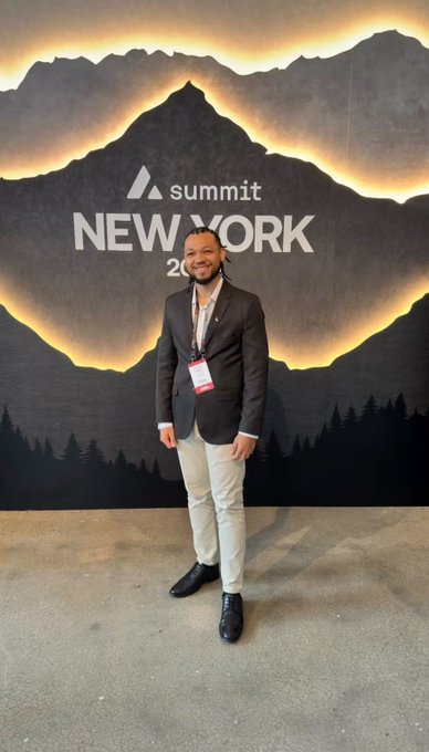 A man in a dark blazer and light pants stands smiling in front of a 'summit NEW YORK' mountain-themed backdrop, wearing a lanyard.