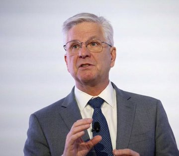A man with gray hair and glasses wearing a gray suit, white shirt, and blue tie appears to be speaking, possibly at a corporate event.