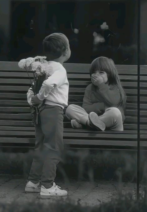 A black-and-white photograph of a young boy holding flowers while facing a girl seated on a bench with her hand over her mouth.