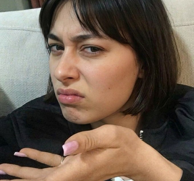 A close-up of a woman with short dark hair and a pouting expression resting her chin near her hands, wearing a black top.