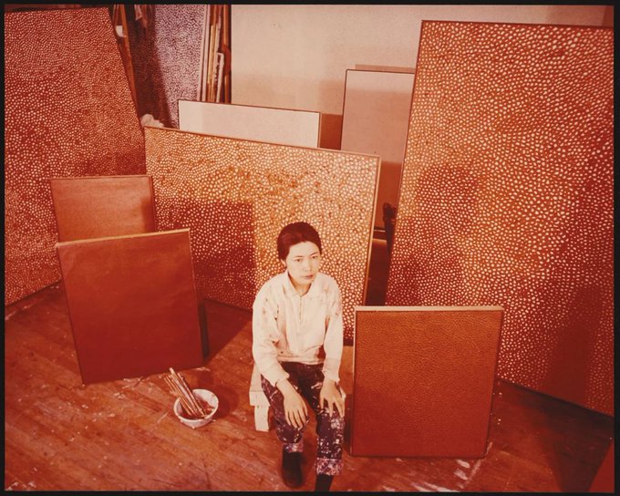 A vintage-style sepia photograph of a woman sitting in an artist's studio surrounded by large canvases covered in orange polka dots, with paintbrushes nearby.