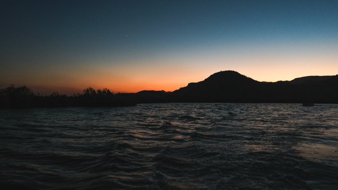 A serene twilight seascape captures the sun setting behind a silhouetted mountain range, with rippling dark waters in the foreground reflecting the fading orange glow on the horizon, evoking a sense of quiet romance and longing that mirrors the post's poetic tribute to sunsets, the sea, and enduring love.