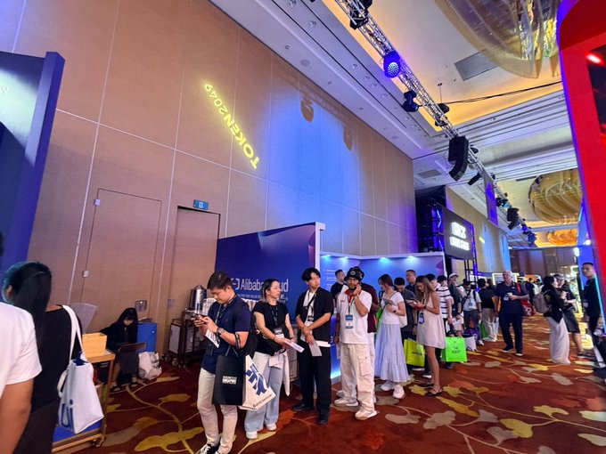 Crowds of attendees queue at the HTX Ventures booth at the TOKEN2049 venue, where staff hand out branded tote bags during a busy giveaway event. Visible signage includes "TOKEN2049" on the back wall and partial branding reading "Alibaba..." alongside an event app banner.

The image directly reflects the post's description of the HTX Ventures booth's overwhelming popularity, showing a steady line of visitors waiting to participate in the giveaway activity.