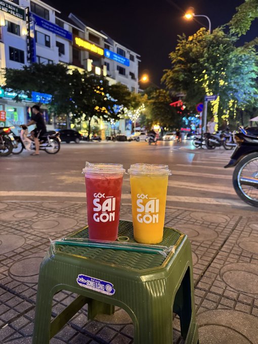 Two plastic cups of 'Sai Gon' beverages—one red and one orange—sitting on a small green plastic stool on a nighttime city sidewalk.