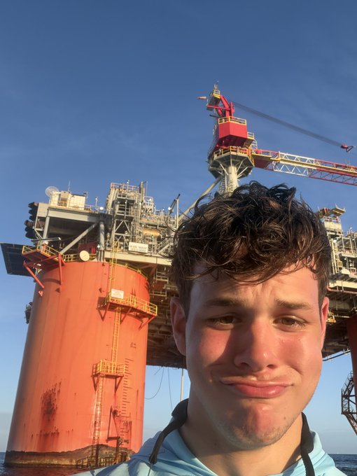 The image features a young man with curly hair posing for a photo near an oil rig. He is wearing a blue shirt, which contrasts with his dark hair. The oil rig behind him has several levels of pipes and equipment, indicating its industrial nature. The caption on the image reads "OUT HERE SHILLING $TROLL AT THE OIL RIGS", suggesting that the man might be involved in an oil-related activity or job at the rig.