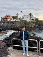 The image features a man standing near a railing overlooking the ocean. The man appears to be wearing a jacket and jeans while posing for the photo. In the background, there are buildings that seem to be older or have a more traditional design compared to the modern cityscape in the foreground. The buildings appear to be made of stone, giving them an old-world charm. The man's position near the railing suggests he is taking a moment to appreciate the view and possibly contemplate his life choices.