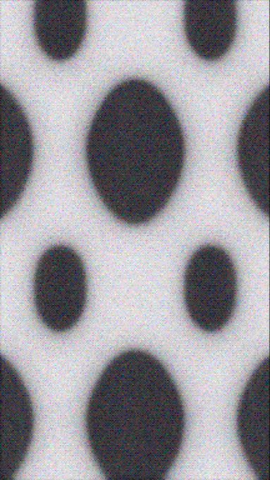 A grayscale image showing a pattern of dark, blurred oval shapes arranged on a lighter, textured background.