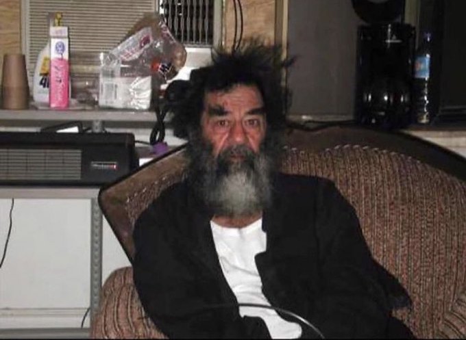 A photograph of a disheveled man with a long gray beard and messy hair, wearing a black jacket over a white shirt, sitting in an armchair in what appears to be a cluttered indoor setting.