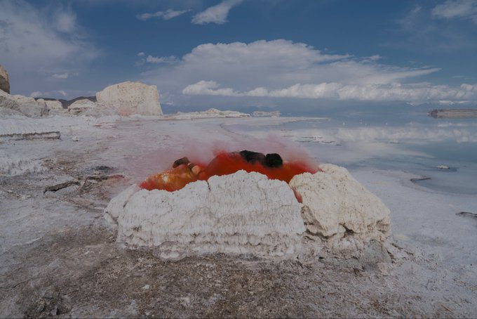 A landscape photograph showing a white, mineral-encrusted formation with a striking red and orange liquid pool in its center, set beside a calm blue body of water under a partly cloudy sky.
