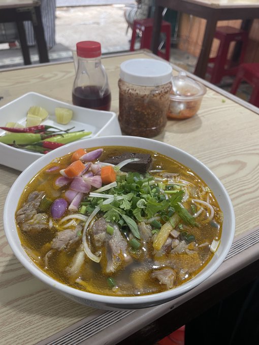 Bowl of Vietnamese beef noodle soup (bún bò) with chili sauce, pickled vegetables, and chili peppers on a wooden restaurant table.