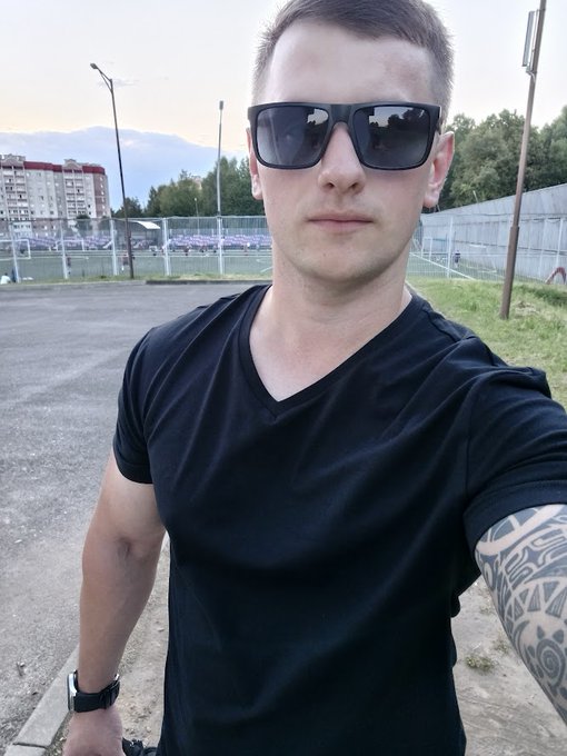 A selfie of a man wearing dark sunglasses and a black v-neck t-shirt, with a tribal tattoo visible on his forearm, standing outdoors with apartment buildings and a fenced sports field in the background.