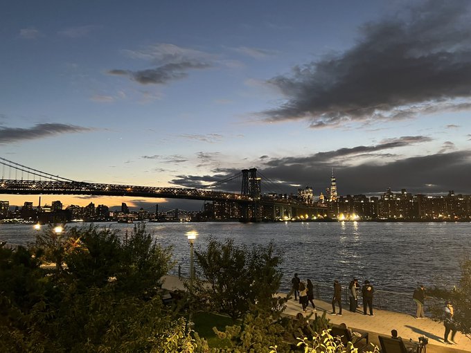 A twilight cityscape showing the Brooklyn Bridge spanning the East River toward Lower Manhattan, with people gathered at a waterfront park in the foreground.