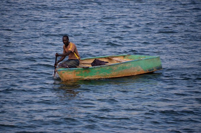 A young boy in an orange and yellow shirt paddling a weathered green and yellow wooden boat across calm blue water.