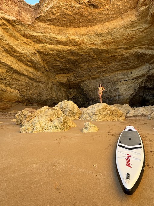 A photograph of a beach inside a cave-like rock formation, with a person standing on the rocks and a white paddleboard lying on the sand in the foreground.