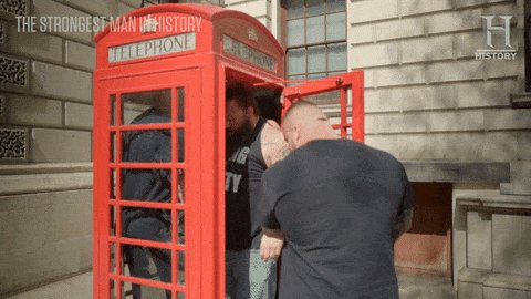 The image features a red telephone booth with three men standing inside, one of whom has his arm around another man's neck. The background shows a building, suggesting that they are located in an urban setting. The caption reads "red phone booth Miami speakeasy October @BoredApeYC today's conspiracy thoughts". This implies that the image is related to a post about a possible conspiracy or unusual event involving the telephone booth and its occupants.