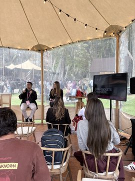 Two people seated on stools under a tent/canopy at an outdoor event, with an audience in front of them and a screen displaying what appears to be the Fauda TV show logo.