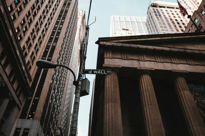 A low-angle photograph of tall buildings in a financial district, with a street sign reading 'WALL ST' attached to a lamppost in the foreground.