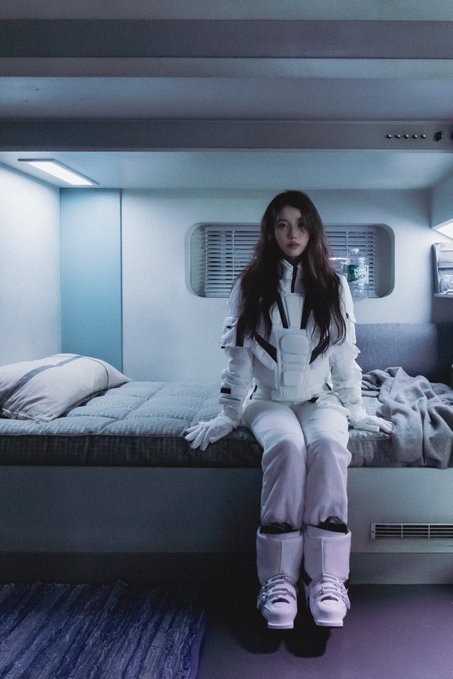 A person with long dark hair sits on a bed inside a futuristic, blue-lit pod-style room, wearing a white tactical vest and chunky white platform boots.