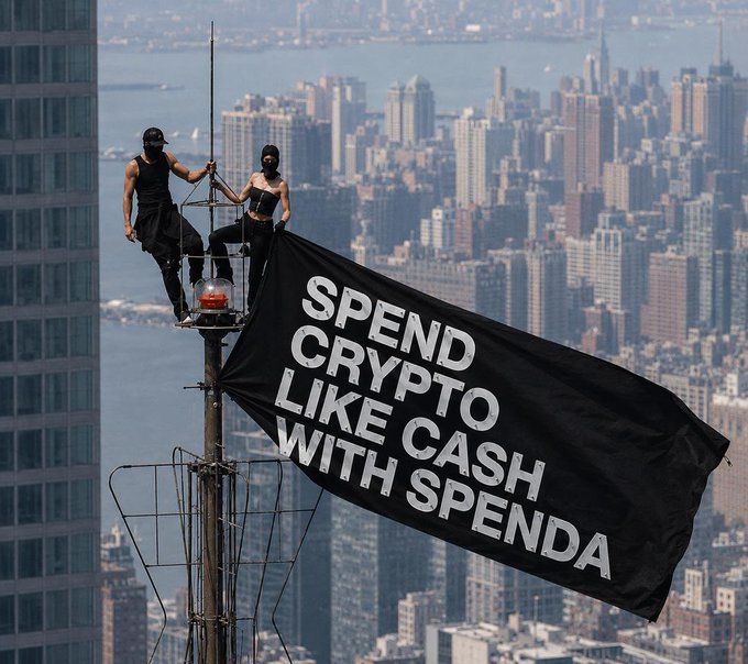 Two people in black outfits stand on a tall structure atop a city skyscraper, holding a large black flag that reads 'SPEND CRYPTO LIKE CASH WITH SPENDA' against a New York City skyline.