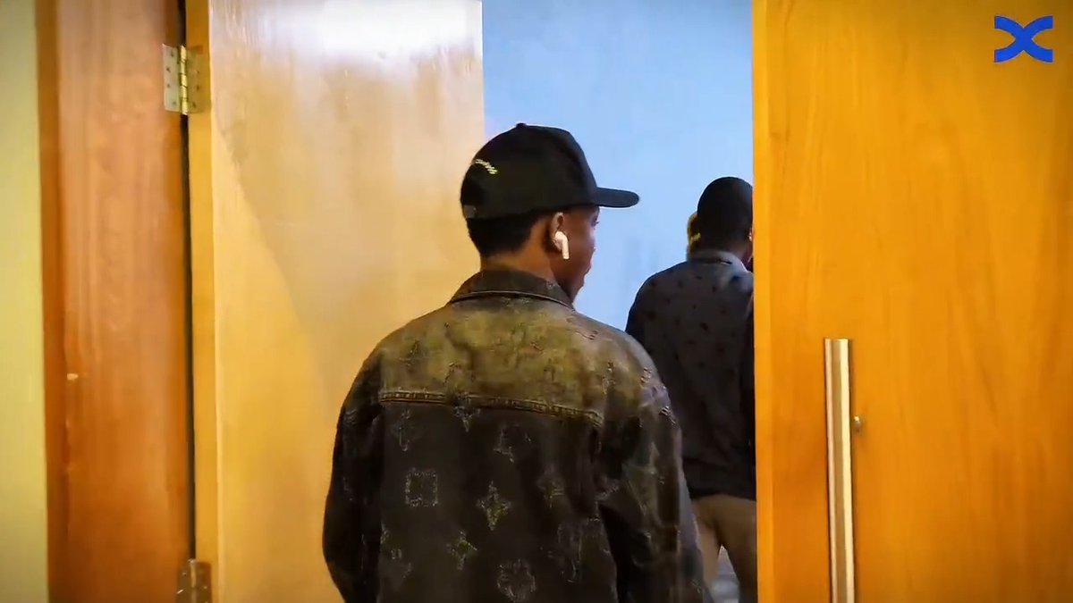 Two attendees—one in a denim jacket and black cap with an earbud, the other in a patterned shirt—walk through wooden doors into a room, capturing the behind-the-scenes arrival of participants at the BingX Kenya Connect 2026 event referenced in the post.
