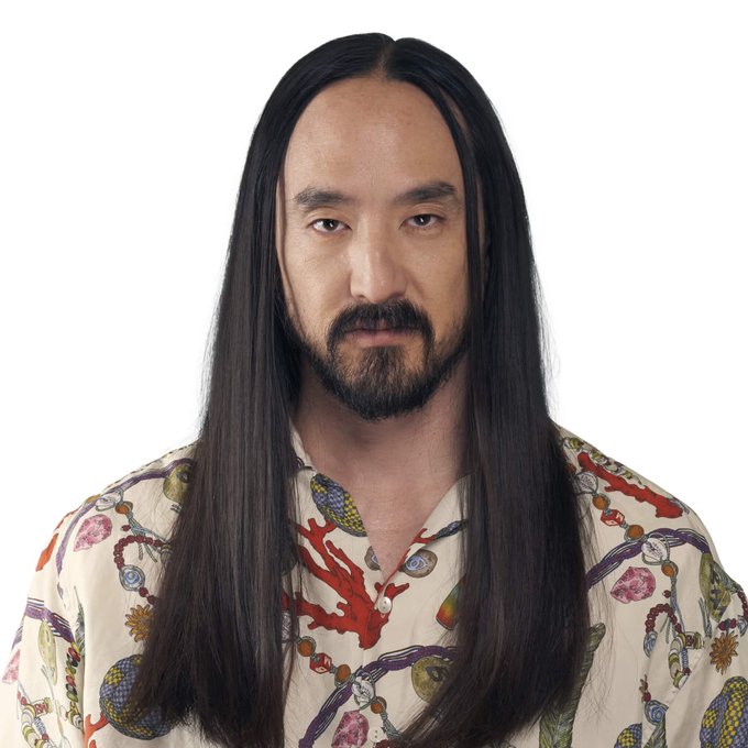 A man with long straight black hair, a beard, and mustache wears a colorful patterned shirt featuring coral and gem designs against a white background.
