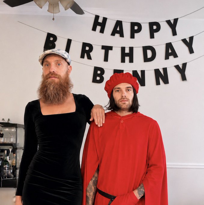 Two men posing at a birthday party in front of a 'HAPPY BIRTHDAY BUNNY' banner; one wears a black dress with a cap and beard, while the other wears a red hooded robe.