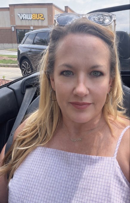 A woman with blonde hair and sunglasses perched on her head takes a selfie in a convertible car. Behind her, a Subway restaurant sign is visible, along with a parked SUV in the lot.