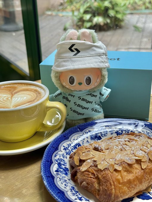 A plush toy wearing a 'Bitget' branded hoodie and visor sitting on a table next to a latte, an almond croissant, and a Bitget gift box.