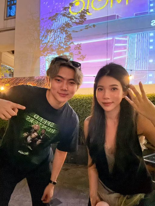 A nighttime selfie of a young Asian couple posing together, with the man pointing at his 'Pirates of the Caribbean 2: Dead Man's Chest' t-shirt, against a background of neon purple lighting.