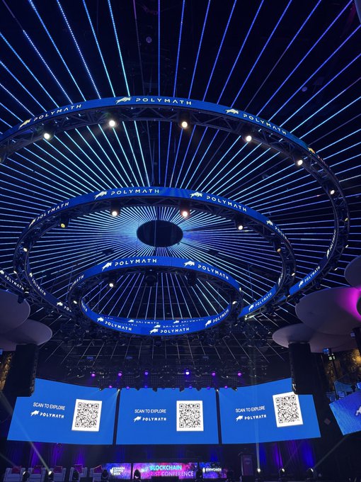 A conference venue features an elaborate circular lighting installation with blue LED rays radiating outward, surrounded by screens displaying 'POLYMATH' logos and QR codes for exploration.