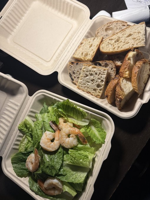 Two takeout containers are open on a dark surface; one holds a shrimp Caesar salad with romaine lettuce, and the other contains an assortment of sliced artisan bread.