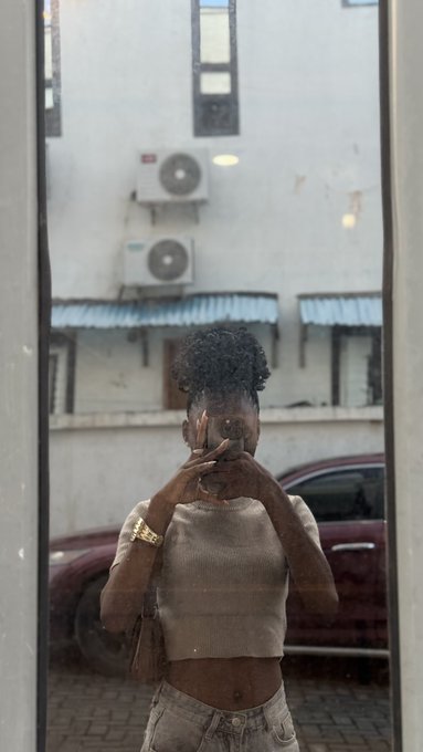 A mirror selfie of a woman with curly hair pulled up, wearing a beige ribbed top and jeans, with her face partially obscured by her phone, standing in front of a building with air conditioning units.