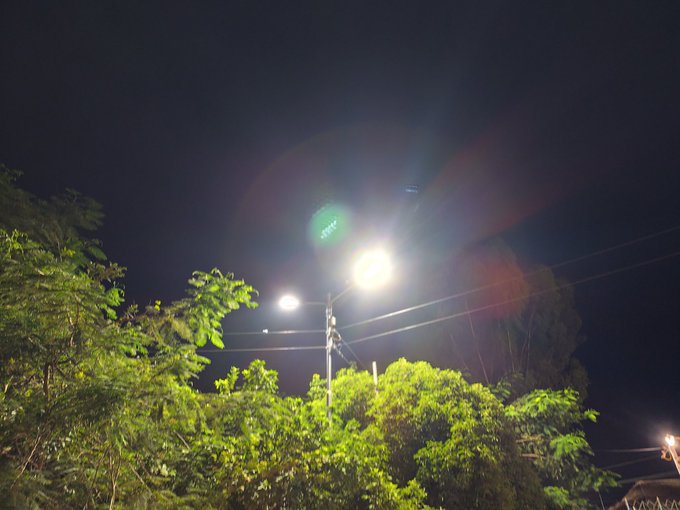 The image features a street lamp with three lights shining brightly against the dark sky at night. The scene includes trees, bushes, and power lines that stretch across the frame. The photo has been edited to emphasize the brightness of the street light, creating an interesting contrast between the illuminated area and the surrounding darkness.