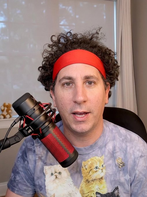 A man with curly hair and a red headband speaking into a red and black microphone, wearing a tie-dye t-shirt featuring kittens with a glitch effect.