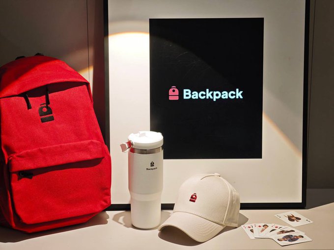 A product display featuring a red backpack, white tumbler, and beige cap branded with the 'Backpack' logo, alongside a framed poster and small trading cards.