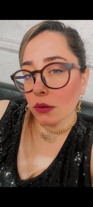 A stylish woman with glasses, red lipstick, gold chain necklace, and a black sequined top poses for a selfie, likely promoting her "Memory Fisher" NFT collection on objkt.com. The caption references the 13/15 edition of the piece, priced at 16 XTZ, with a playful nod to her collaborator @King_PRomulus.