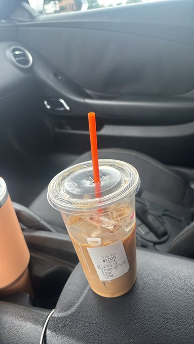 An iced coffee in a clear plastic cup with an orange straw sits in a car's center console next to a tumbler.