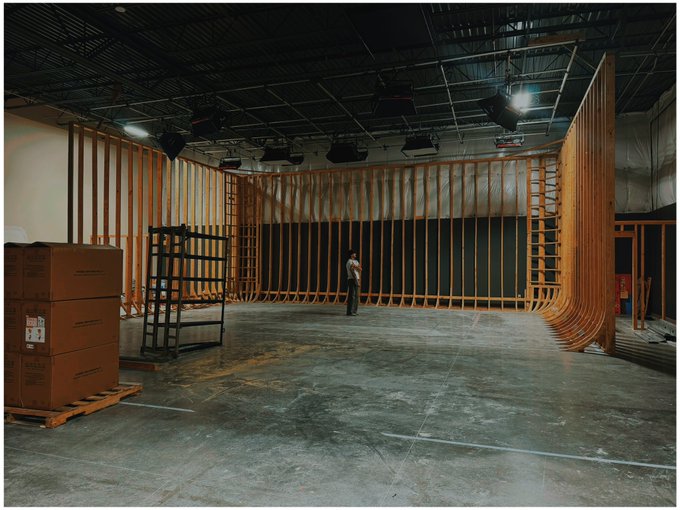A large warehouse or studio space under construction, featuring wooden framing, cardboard boxes, and a person standing in the center.
