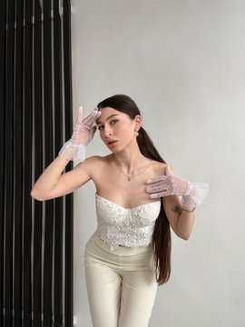 A young woman poses indoors in an elegant white strapless corset top and cream-colored pants, wearing sheer gloves and with long brown hair, touching her face against a dark paneled background.