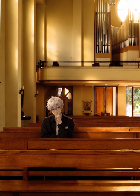 An anime-styled character with silver hair and glasses sits alone in a wooden church pew, resting their chin thoughtfully on their hand beneath warm, golden lighting.