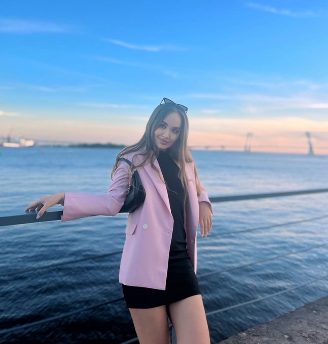 A woman in a pink blazer and black dress leans against a railing by a waterfront, with a bridge visible in the background at sunset.