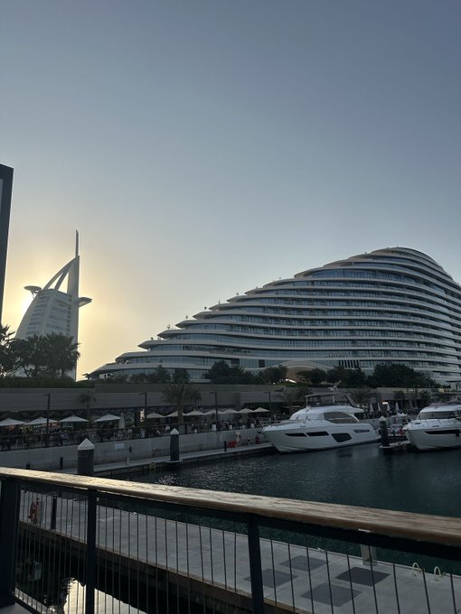 A scenic sunset view of Dubai Marina featuring the iconic Burj Al Arab hotel in the distance and yachts docked along the waterfront.