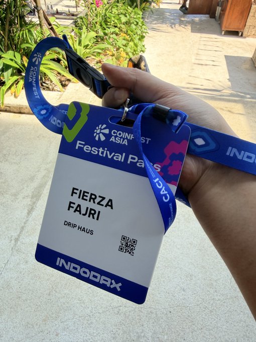 A hand holds a blue lanyard with a 'Coinfest Asia' festival pass for 'Fierza Fajri' from 'Drip Haus', also displaying an 'INDORAX' logo and QR code.